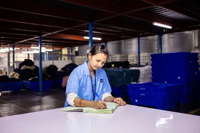 Woman in blue uniform taking notes in an industrial warehouse setting.